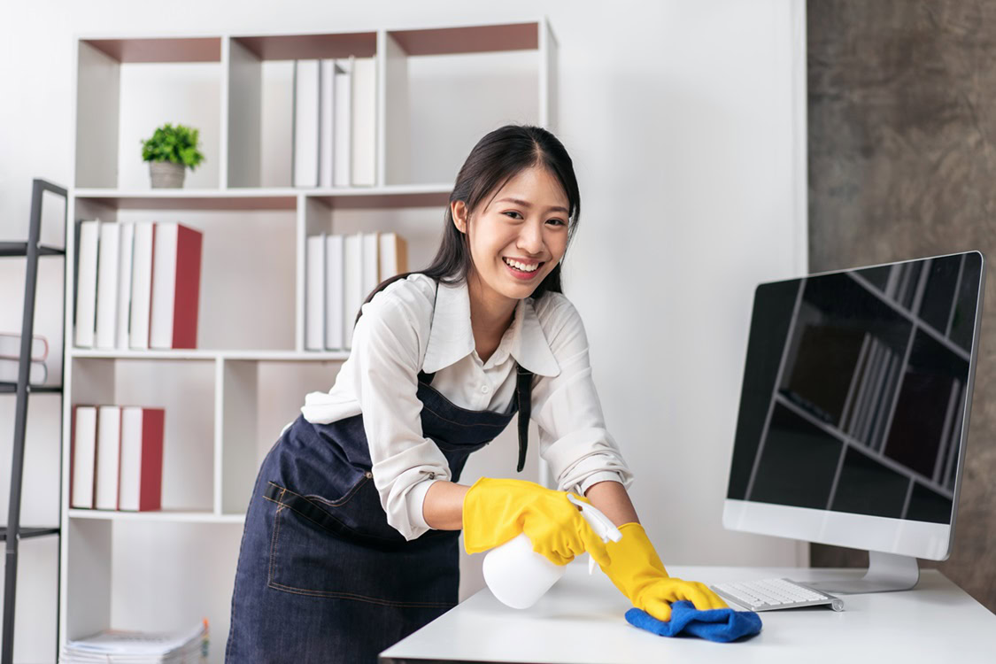 A person wearing gloves, smiling as they clean a desk with a spray bottle and cloth, embodies the professional touch of Cleaning Services in Kansas City. In the background, neatly organized bookshelves and a computer monitor reflect the gleaming results of their meticulous care.