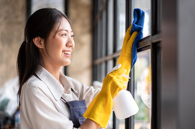 A person in an apron, representing top-notch cleaning services in Kansas City, meticulously cleans a window with a blue cloth and spray bottle, donning yellow rubber gloves for protection.