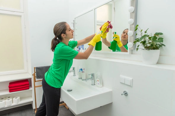 A woman in a green shirt and yellow gloves cleans a bathroom mirror with precision, representing the quality you can expect from cleaning services in Kansas City. A white sink gleams below, while a green plant adds a touch of nature to the spotless space.