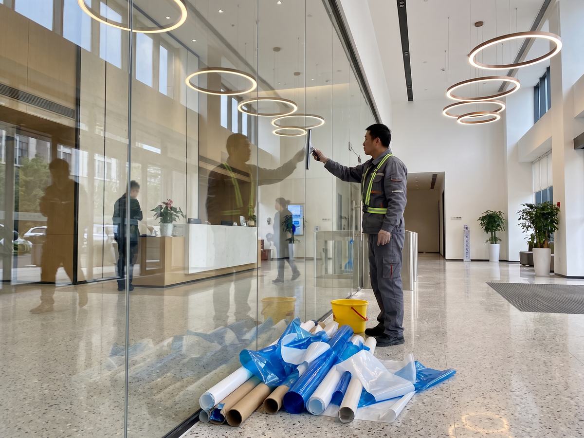 Cleaning technician squeegeeing a new glass partition in a commercial lobby during touch-up cleaning, with peeled protective film stacked on the floor